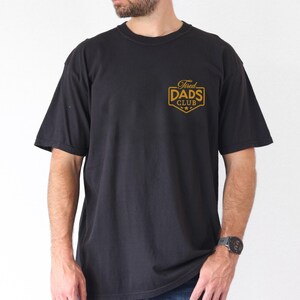 May include: Black short-sleeved t-shirt with the text "Tired Dads Club" in gold lettering on the chest. The shirt is a classic crew neck style and appears to be made of a soft, comfortable material.