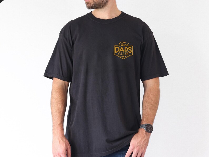 May include: Black short-sleeved t-shirt with the text "Tired Dads Club" in gold lettering on the chest. The shirt is a classic crew neck style and appears to be made of a soft, comfortable material.