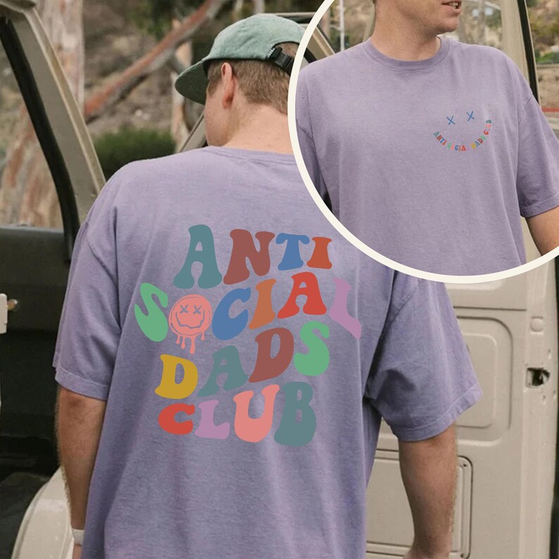May include: A lilac-colored t-shirt with the text "ANTI SOCIAL DADS CLUB" in a retro font on the back. The front of the shirt features a small smiley face design with the words "ANTI SOCIAL CLUB" in a circular pattern. The shirt has short sleeves.