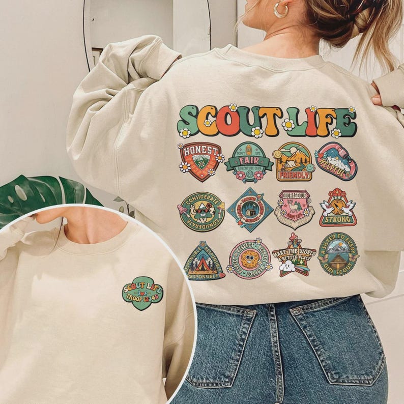 Puede incluir: Sudadera beige con el texto "SCOUT LIFE" en letras coloridas y detalles florales. La parte trasera de la sudadera presenta varios parches con palabras como "HONEST", "FAIR" y "STRONG". Una versi&oacute;n m&aacute;s peque&ntilde;a del dise&ntilde;o est&aacute; en la parte delantera.