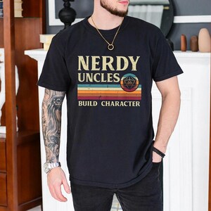 May include: A black short-sleeved t-shirt featuring the text NERDY UNCLES BUILD CHARACTER in a vintage font. The design includes a multi-colored horizontal striped graphic and a circular icon containing a twenty-sided die symbol in the center.