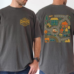 May include: Dark gray t-shirt with the text "Tired Dads Club" on the front and a colorful road map design on the back. The back design includes houses, cars, and a tractor.