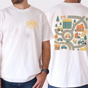 May include: White t-shirt with a graphic on the front that says "Proud Dads Club" in a yellow badge. The back of the shirt features a colorful illustration of a town with roads, houses, trees, and vehicles like a tractor and ambulance.