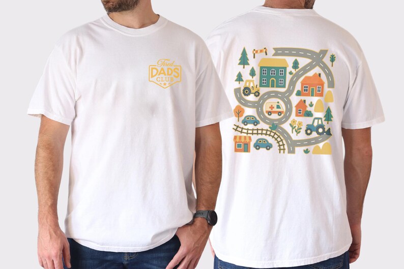 May include: White t-shirt with a graphic on the front that says "Proud Dads Club" in a yellow badge. The back of the shirt features a colorful illustration of a town with roads, houses, trees, and vehicles like a tractor and ambulance.