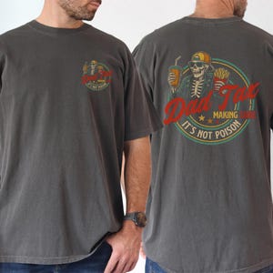 Pu&ograve; includere: T-shirt grigio scuro con grafica sul davanti e sul retro. La grafica anteriore &egrave; un design circolare con testo. La grafica posteriore presenta uno scheletro con cappello e occhiali da sole, che tiene in mano una bevanda e patatine fritte, con la scritta "Dad Fuel, It's Not Poison."