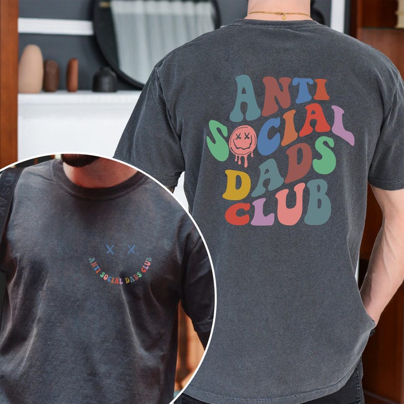 May include: Dark gray t-shirt with the text "ANTI SOCIAL DADS CLUB" in colorful, retro-style lettering on the back. The front of the shirt features a smiling face with crossed-out eyes and the same text in a semi-circle. A casual, comfortable tee.