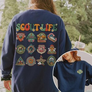 Puede incluir: Sudadera azul marino con la frase "SCOUT LIFE" en letras retro coloridas. Debajo, varios parches coloridos con palabras como "Honest" y "Fair". Una versi&oacute;n m&aacute;s peque&ntilde;a del dise&ntilde;o se encuentra en la parte inferior derecha.