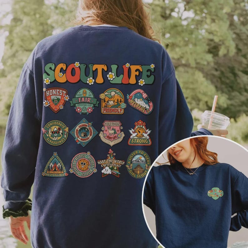 Puede incluir: Sudadera azul marino con la frase "SCOUT LIFE" en letras retro coloridas. Debajo, varios parches coloridos con palabras como "Honest" y "Fair". Una versi&oacute;n m&aacute;s peque&ntilde;a del dise&ntilde;o se encuentra en la parte inferior derecha.