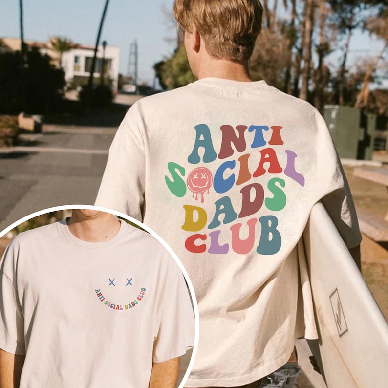 May include: Cream-colored t-shirt with the text "ANTI SOCIAL DADS CLUB" in colorful, retro-style lettering. The design includes a smiley face with crossed-out eyes. The shirt is a casual, oversized fit.