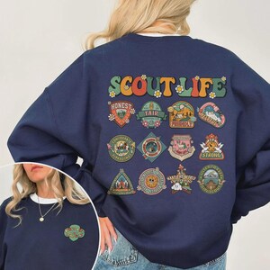 Puede incluir: Sudadera azul marino con la frase "SCOUT LIFE" en letras retro coloridas. La parte trasera de la sudadera presenta m&uacute;ltiples parches con palabras como "HONEST", "FAIR" y "STRONG". Una versi&oacute;n m&aacute;s peque&ntilde;a del dise&ntilde;o est&aacute; en la parte delantera.