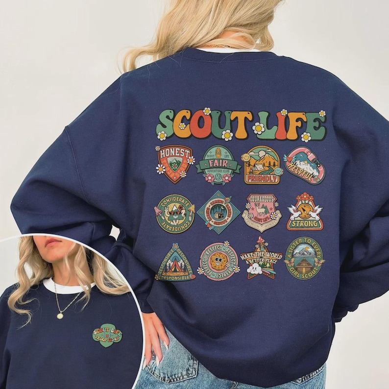 Puede incluir: Sudadera azul marino con la frase "SCOUT LIFE" en letras retro coloridas. La parte trasera de la sudadera presenta m&uacute;ltiples parches con palabras como "HONEST", "FAIR" y "STRONG". Una versi&oacute;n m&aacute;s peque&ntilde;a del dise&ntilde;o est&aacute; en la parte delantera.
