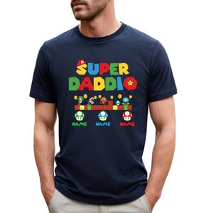 May include: Navy blue t-shirt with a colorful graphic design. The design features the words "SUPER DADDIO" in yellow, green, red, and blue, along with cartoon characters and the word "NAME" below each character.