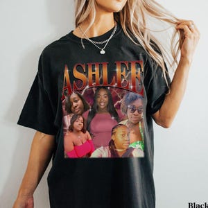 May include: Black t-shirt with a graphic print of the word "ASHLEE" in red and yellow letters with lightning bolts. The graphic also features five women's faces in a collage style.