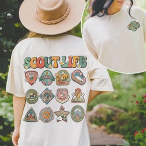 Può includere: T-shirt color crema con la scritta "SCOUT LIFE" in lettere retrò colorate. La parte posteriore della maglietta presenta varie toppe con parole come "HONEST", "FAIR" e "FRIENDLY". Una versione più piccola del disegno è sul davanti.