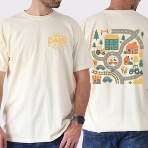 May include: Cream-colored t-shirt with a graphic on the back featuring a road map design with houses, vehicles, and trees. The front of the shirt has a gold-colored "Dads Club" logo. The shirt is made of a soft, comfortable material.
