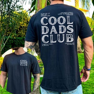 Peut inclure: T-shirts noirs avec l'inscription blanche "COOL DADS CLUB". L'un des t-shirts porte le texte sur le devant, l'autre au dos avec les phrases: "ALWAYS UP FOR AN ADVENTURE", "UNCONDITIONAL LOVE, ALWAYS AND FOREVER", "FAMILY FIRST, ALWAYS", et "LEADING BY EXAMPLE".