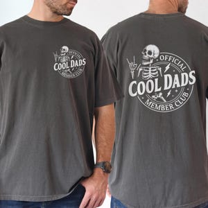 Pode incluir: T-shirt cinzento escuro com o texto "OFFICIAL COOL DADS MEMBER CLUB" e um gráfico de esqueleto. O desenho é impresso na frente e nas costas. A t-shirt tem gola redonda e mangas curtas.