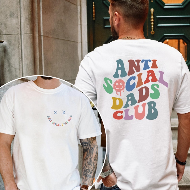 May include: White t-shirts with the text "ANTI SOCIAL DADS CLUB" in colorful, retro-style lettering. One shirt features the text on the back, and the other has a smiley face and the text on the front. The shirts are a casual style.