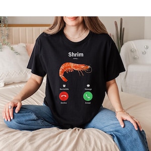May include: Black t-shirt featuring a shrimp graphic and the word "Shrim" in white. The design resembles a phone screen, with "calling" above the shrimp image and "Decline" and "Accept" buttons below.