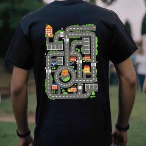 May include: A black t-shirt with a colorful graphic of a city street design. The design features a circular road with houses, trees, and cars.