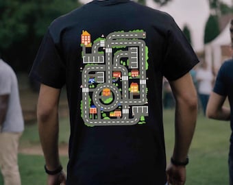 Può includere: Una maglietta nera con una grafica colorata di un design di strada cittadina. Il design presenta una strada circolare con case, alberi e auto.