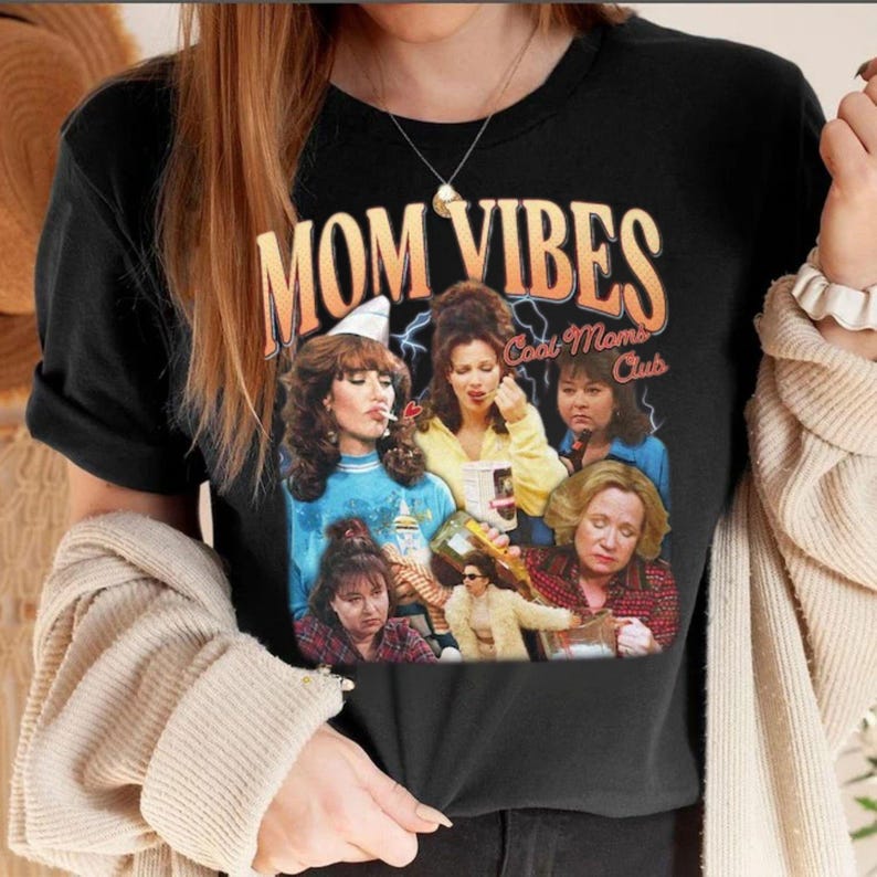 May include: Black t-shirt with a vintage-style graphic featuring a group of women and the text "Mom Vibes Cool Moms Club".