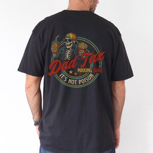 Pu&ograve; includere: T-shirt nero con grafica sul retro raffigurante uno scheletro con cappello e occhiali da sole, che tiene in mano una bevanda e patatine fritte. Il testo recita "Dad Tax MAKING SURE IT'S NOT POISON" in un design circolare.