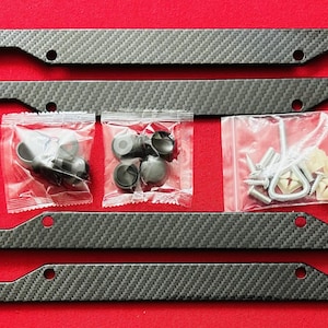 May include: A black carbon fiber license plate frame with two sets of mounting hardware. The hardware includes black plastic push-in fasteners, silver metal screws, and silver metal hooks.