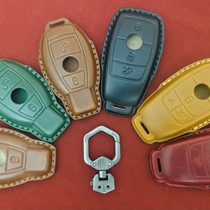 May include: A collection of leather key fob covers in various colors, including green, brown, black, and yellow, with white stitching. A silver-colored metal carabiner is also present. The key fobs are arranged on a red surface.