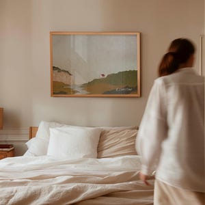 May include: A white linen bedding set with a beige throw pillow on a bed with a wooden headboard. A framed landscape painting hangs on the wall behind the bed. A person is walking towards the bed.
