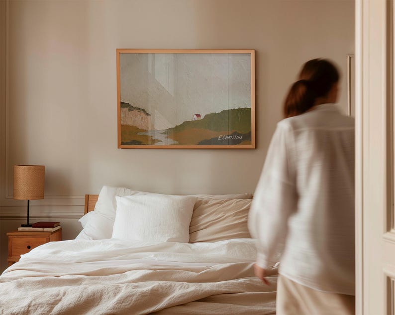 May include: A white linen bedding set with a beige throw pillow on a bed with a wooden headboard. A framed landscape painting hangs on the wall behind the bed. A person is walking towards the bed.