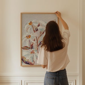 May include: A person hangs a framed painting of white flowers on a white wall. The painting is in a light wood frame and the flowers are painted in a loose, impressionistic style.