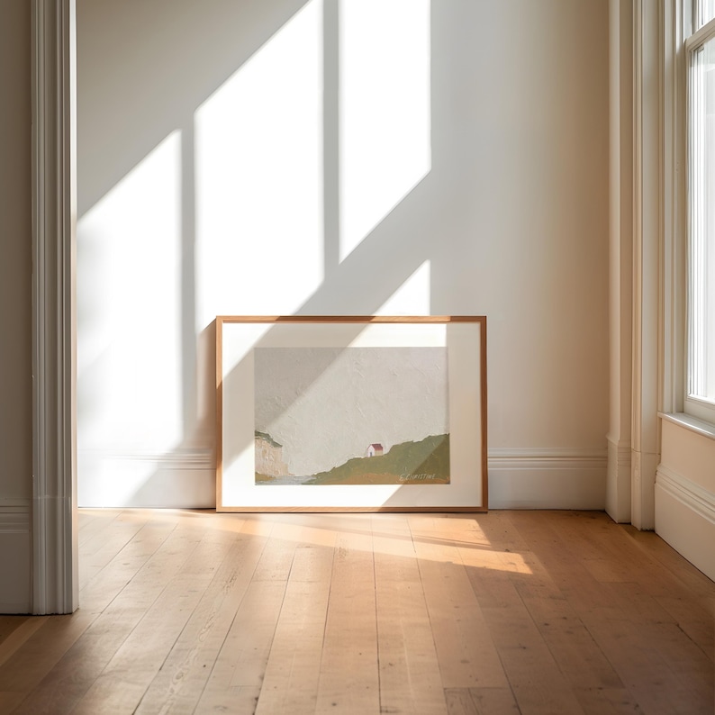 May include: A framed landscape painting of a green hillside with a small white house and a cliff in the distance. The painting is in a light wood frame and is hanging on a white wall.