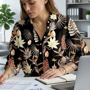 Può includere: Camicia nera con bottoni con stampa botanica con foglie e fiori beige, arancioni e gialli. La camicia è indossata da una persona che lavora a una scrivania con un laptop e documenti.