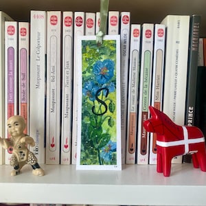 Peut inclure: Marque-page rectangulaire à l'aquarelle avec des fleurs bleues et des feuilles vertes, et une lettre noire "S". Un ruban vert est attaché en haut. Le marque-page est devant une bibliothèque.