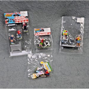 May include: Several miniature figurines in clear plastic packaging. One package contains six figurines of soccer players. Another package has two figurines, one of a person and a cow. A third package has an ice cream cart and a person. A small bag contains additional figurines.