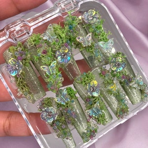 Op de afbeelding: Een set van tien groene glitter press-on nagels met 3D vlinder- en bloemenversieringen. De nagels zijn gerangschikt in een doorzichtige plastic doos.