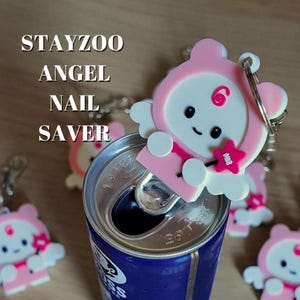 May include: A pink and white Stayzoo Angel Nail Saver keychain is perched on top of a blue and silver can. The keychain features a cartoon angel design with a wand. The text "STAYZOO ANGEL NAIL SAVER" is visible.