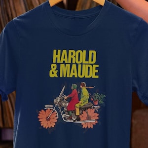 Puede incluir: Una camiseta azul marino con un gráfico amarillo del texto "HAROLD & MAUDE" y una ilustración vintage de dos personas en una motocicleta.