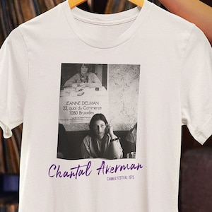 Puede incluir: Camiseta blanca con una foto en blanco y negro de una mujer y el texto "Jeanne Dielman 23, quai du Commerce 1080 Bruxelles" y "Chantal Akerman Cannes Festival 1975".