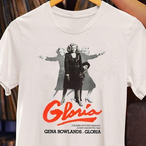 May include: White t-shirt with a black and white image of a woman in a black dress and a red title that says "Gloria". The text below the image says "Columbia Pictures Presents A John Cassavetes Film Gena Rowlands in Gloria".