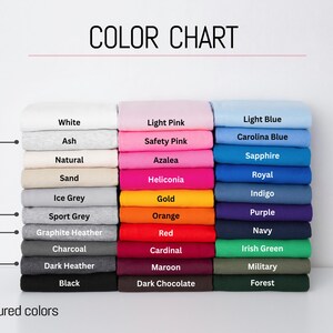 May include: A color chart displaying three vertical stacks of folded fabric in various shades. The labels include White, Ash, Natural, Sand, Ice Grey, Sport Grey, Graphite Heather, Charcoal, Dark Heather, Black, Light Pink, Safety Pink, Azalea, Heliconia, Gold, Orange, Red, Cardinal, Maroon, Dark Chocolate, Light Blue, Carolina Blue, Sapphire, Royal, Indigo, Purple, Navy, Irish Green, Military, and Forest.