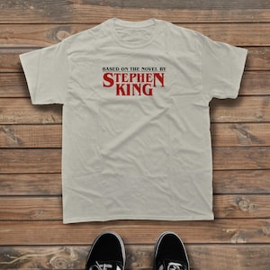 Könnte beinhalten: Ein hellbeiges Kurzarm-T-Shirt mit dem Text "BASED ON THE NOVEL BY STEPHEN KING", der in schwarzen und roten Buchstaben auf der Brust gedruckt ist. Das Shirt hat einen Rundhalsausschnitt und eine Standardpassform, flach auf Holz liegend.