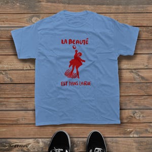 May include: A light blue short-sleeve t-shirt featuring a red graphic of a person jumping above a net. The text "LA BEAUT&Eacute;" is printed above the figure, and "EST DANS LA RUE" is printed below the figure in a bold red font.