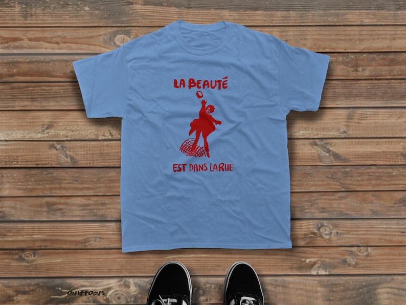 May include: A light blue short-sleeve t-shirt featuring a red graphic of a person jumping above a net. The text "LA BEAUT&Eacute;" is printed above the figure, and "EST DANS LA RUE" is printed below the figure in a bold red font.