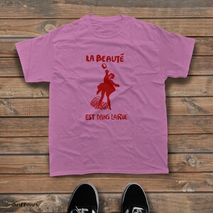 May include: A light pink short-sleeved t-shirt featuring a red graphic of a person jumping above a net. The text "LA BEAUT&Eacute;" is printed in red above the figure, and the text "EST DANS LA RUE" is printed in red below the figure.