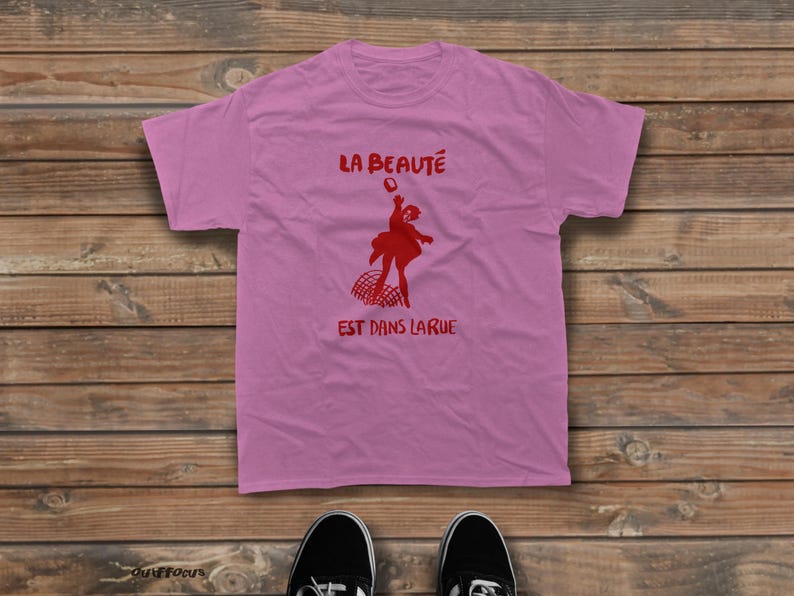 May include: A light pink short-sleeved t-shirt featuring a red graphic of a person jumping above a net. The text "LA BEAUT&Eacute;" is printed in red above the figure, and the text "EST DANS LA RUE" is printed in red below the figure.