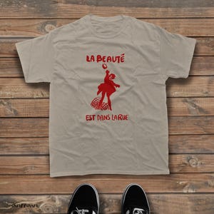 May include: A beige short-sleeved t-shirt featuring a red graphic of a person jumping over a barrier. The text "LA BEAUT&Eacute;" is printed in red above the figure, and "EST DANS LA RUE" is printed in red below the figure on the front of the shirt.