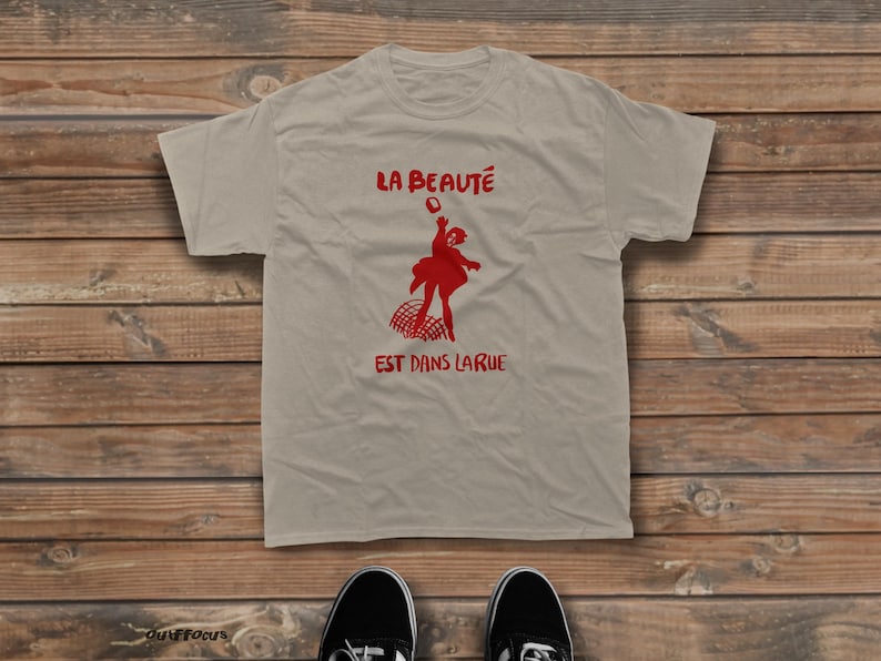 May include: A beige short-sleeved t-shirt featuring a red graphic of a person jumping over a barrier. The text "LA BEAUT&Eacute;" is printed in red above the figure, and "EST DANS LA RUE" is printed in red below the figure on the front of the shirt.