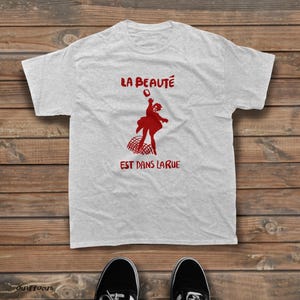 May include: A light gray short-sleeved t-shirt featuring a red graphic print of a person jumping above a net. The text "LA BEAUT&Eacute;" is printed in red above the figure, and "EST DANS LARUE" is printed in red below the figure on the chest.